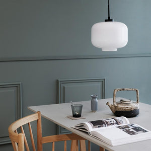 A white glass pendant hangs over a table; below is a rustic teapot, open book, glass and small vase on a bright table surrounded by a dusty blue-grey wall and wooden chair.