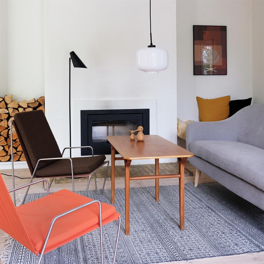 A wooden table stands in the middle of the room and bears two wooden figures; surrounded by gray sofa, orange and brown chair, wood by the fireplace and a hanging lamp in a bright, minimalist room.