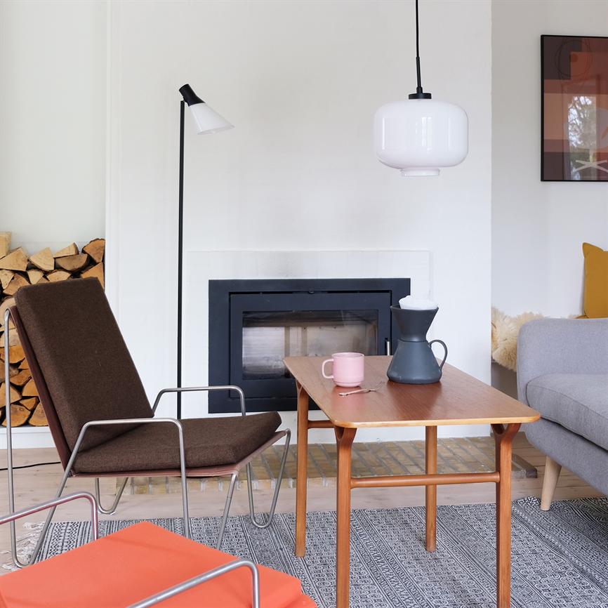 A teak coffee table stands in front of a closed fireplace; a pink mug and a black coffee pot are on the table. Surrounded by armchair, gray sofa, firewood and hanging lamp.