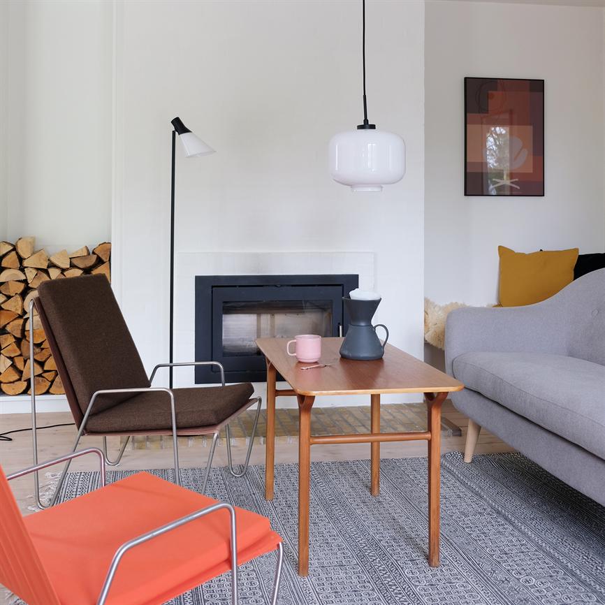 A wooden coffee table stands on a patterned rug in front of a closed fireplace; a dark jug and a pink cup are on the table; modern living room with sofa, chairs and firewood.