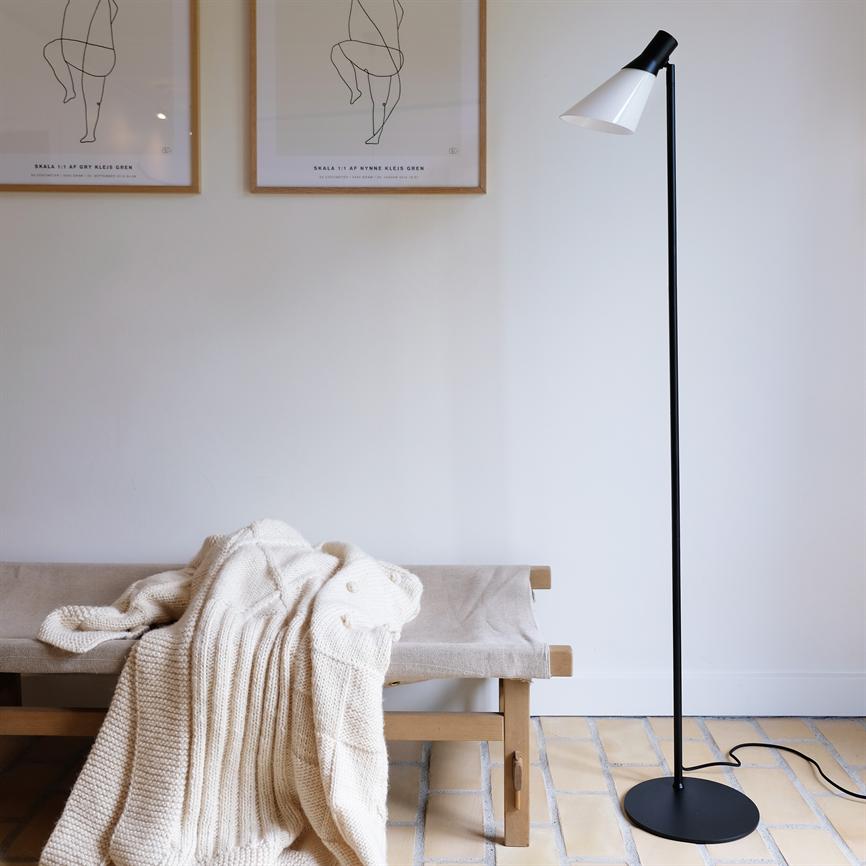 Floor lamp stands to the right by a low wooden bench; a thick, brightly knitted blanket is draped over the bench. White wall with two simple poster drawings. Text on posters: "SCALE 1:1 BY NYNNE KLEIS GREEN".