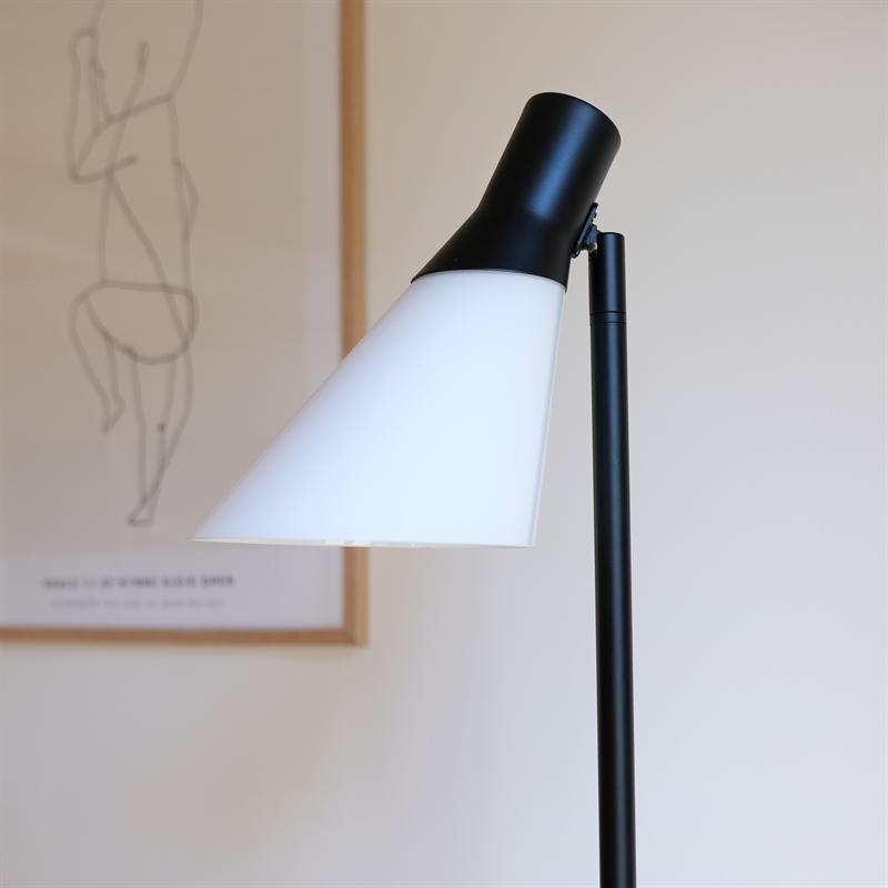 Floor lamp with white cone-shaped shade pointing downwards; black rod and socket. The background shows a blurred line drawing on a light wall in a minimalist room.