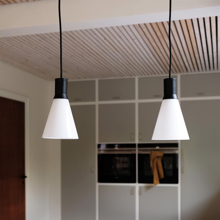 Two white cone-shaped pendant lights hang from black cords above a kitchen, in front of gray built-in cabinets and double ovens; wooden slats in the ceiling and a tea towel hanging on the oven door.