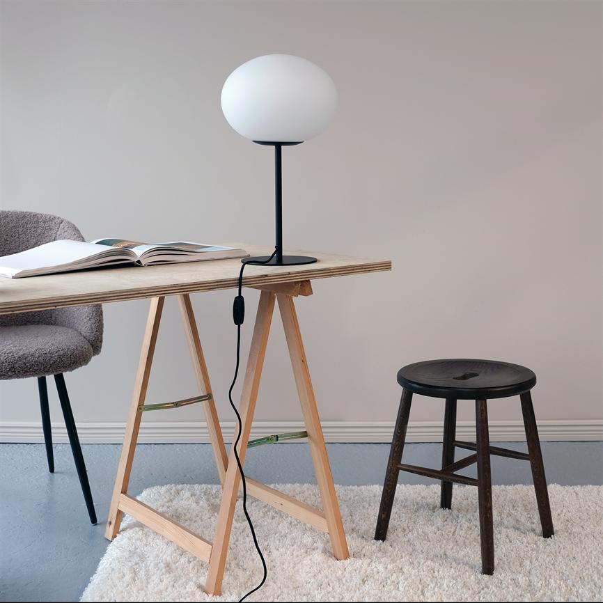 A round table lamp stands on a wooden table next to an open book, surrounded by an upholstered chair, a dark stool and a bright, minimalist room.