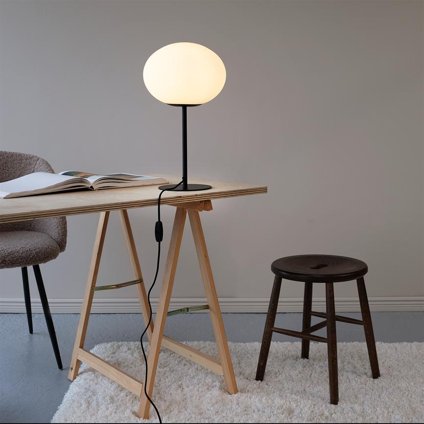 Table lamp glows softly and stands on a simple wooden table with saw legs; an open book, a chair and a dark wooden stool next to it in a calm, minimalist room with light carpet.