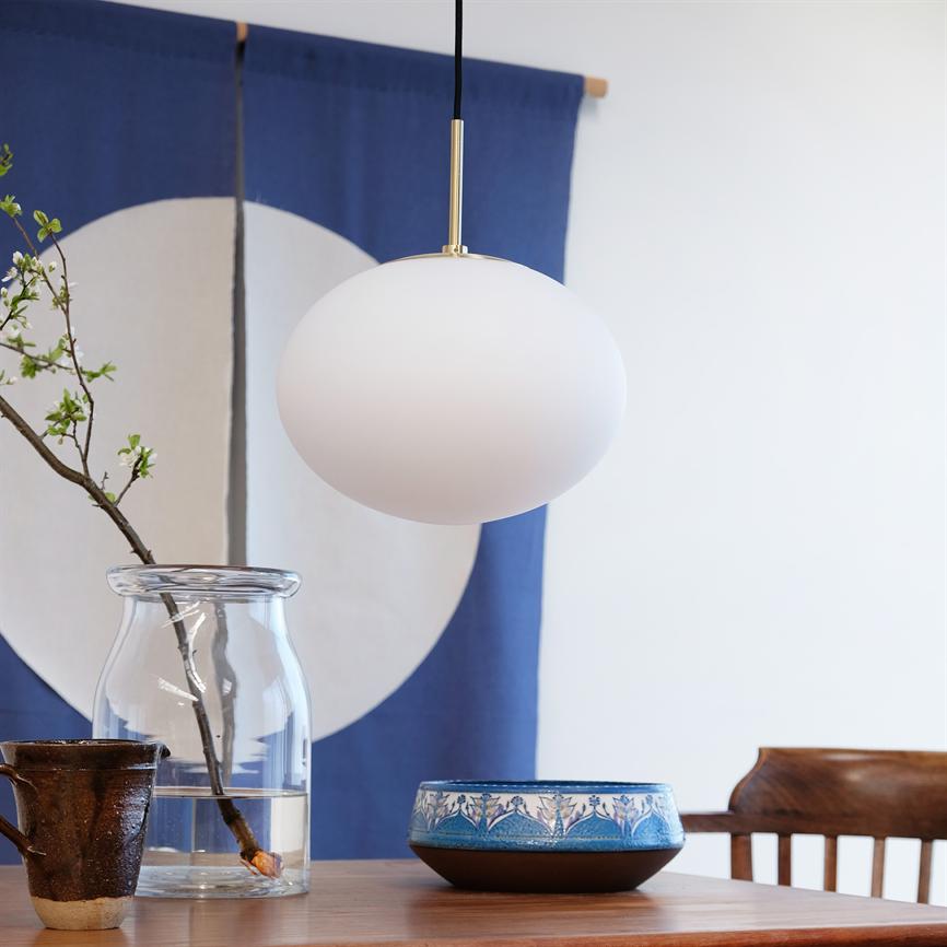 White spherical pendant lamp hangs above a wooden table with a glass vase with a budded sprig, a blue ceramic bowl and a rustic cup, in front of a blue fabric panel with a white circle.