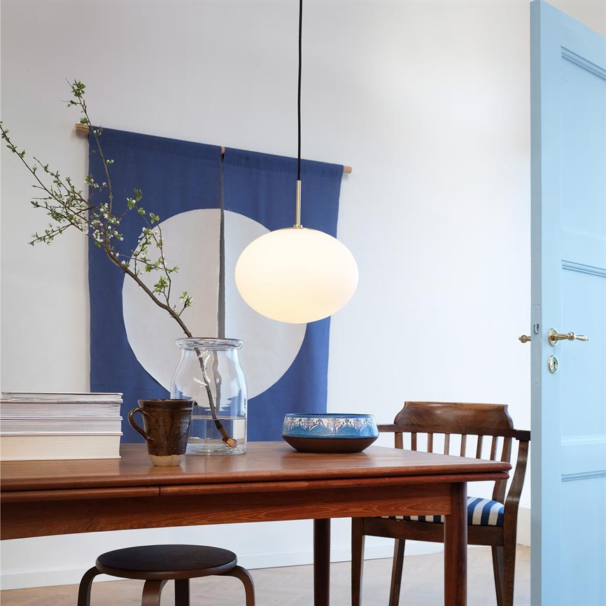 Pendant lamp hangs above a wooden table and casts warm light; on the table is a glass vase with a flowering branch, mug, bowl and books; blue wall textile and open light blue door in the background.