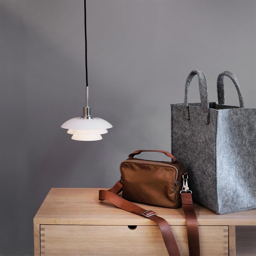pendant hangs and casts soft, warm light over a light wooden chest of drawers, where a brown shoulder bag lies with a strap over the edge and a large gray felt bag stands next to it.