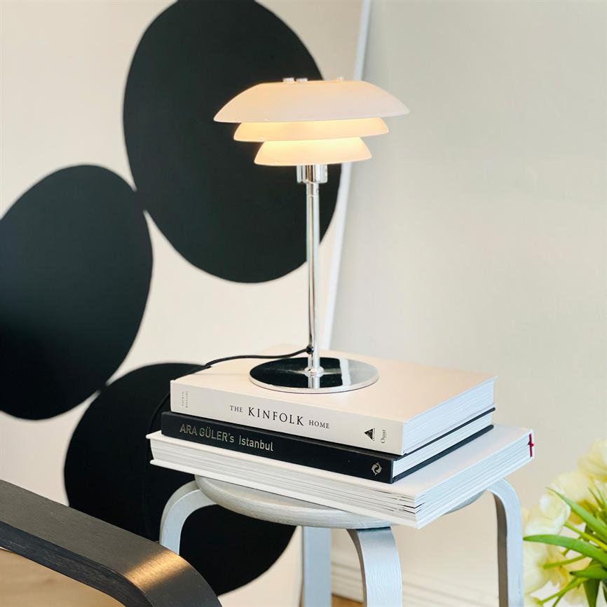 A modern table lamp is lit on a stack of books on a small white stool; black graphic circles on the wall behind and a flower in the lower right corner. 
 Text in the picture: THE KINFOLK HOME; ARA GÜLER's Istanbul