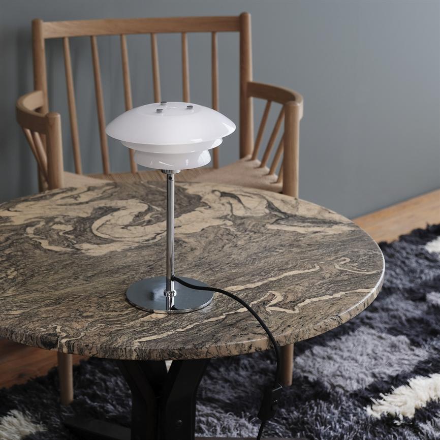 Table lamp stands on a round marble table with beige and dark veins; black wire runs over the edge. Behind a wooden chair against a gray wall and a dark, soft carpet.
