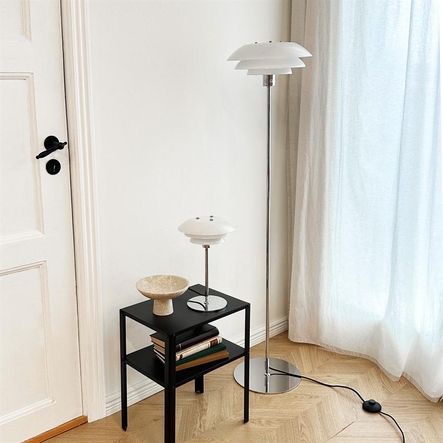 White, layered floor lamp stands by a bright window; a matching table lamp stands on a small black side table with books and a bowl on a light wooden floor by a white door.