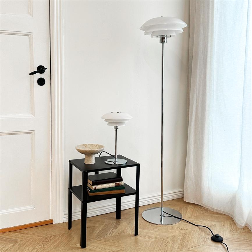 Floor lamp and table lamp stand next to a black side table; the table bears a stone bowl and books. The room is bright with a white wall, door, curtain and wooden floor.