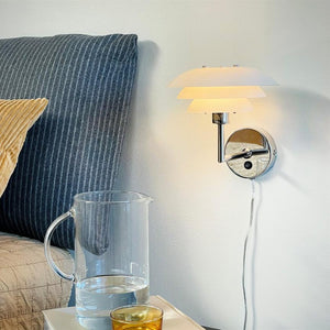 Wall lamp when switched on emits soft warm light; hangs above a bedside table with a half-filled glass jug and a small golden glass, in front of a striped pillowcase and headboard.