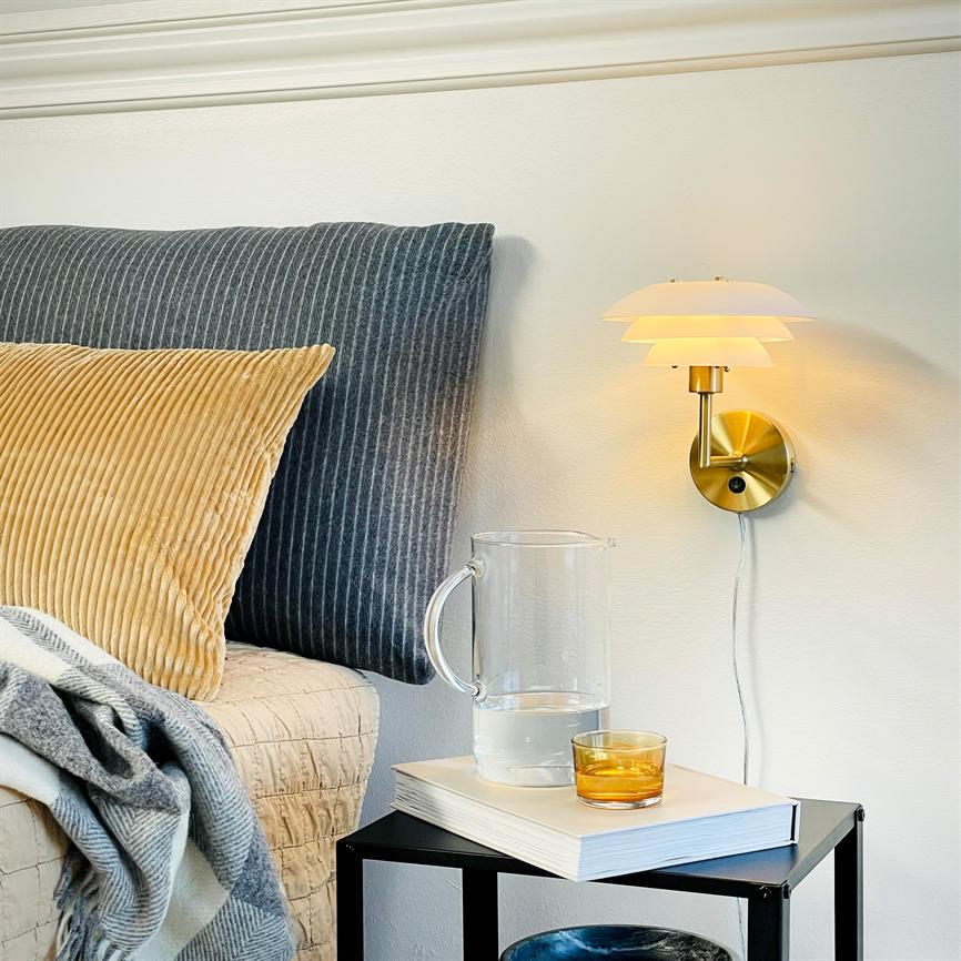 Wall lamp on the wall shines softly above the bedside table; a clear glass decanter and a golden glass stand on a book; striped headboard, beige pillow and blanket in the background.