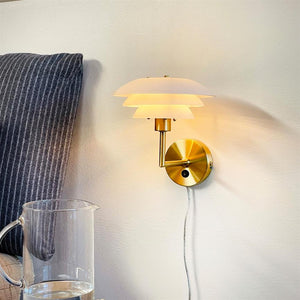 Wall lamp with golden bracket emits warm, diffused light; mounted on a light wall by a striped upholstered headboard, above a bedside table with a glass jug of water.