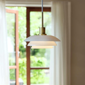 A white multi-layer pendant light hangs lit, casting warm light, in front of open patio doors with white curtains and a blurred view of a green garden beyond.