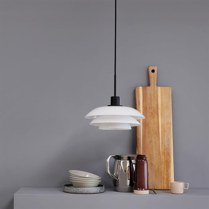 A white, layered pendant lamp hangs above a gray tabletop; next to it is a long cutting board, a silver jug, a brown thermos, four bowls and a small pink cup.