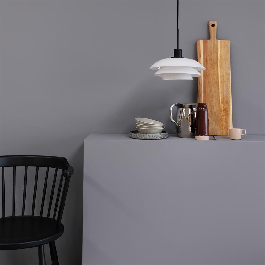 White pendant lamp hangs above a gray sideboard; on the sideboard are bowls, a dish, a blank jug, thermos, a mug and cutting board in front of a gray wall; black chair on the left.