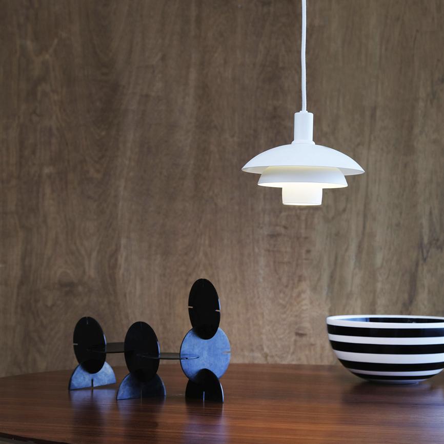 A white one pendant lamp hangs and casts soft light down a wooden table; on the table are three black circular sculptures and a black-and-white striped bowl in front of a warm wood-clad wall.