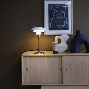 A white table lamp is lit on a light oak sideboard; it casts warm light over a ceramic jar and two sculptural vases beneath a framed, black-and-white abstract poster on a brown wall.