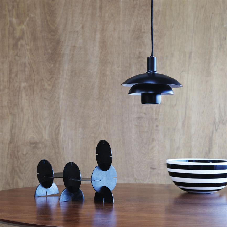 Black pendant hangs over a wooden table; three black, circular sculptures stand on the table, a black-and-white striped bowl to the right; warm wood paneling forms the background.