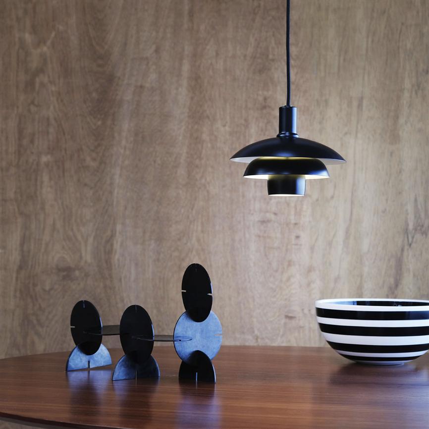 Black metal pendant hangs above a round wooden table; three joined black disc elements stand on the table and a black and white striped bowl stands to the right in front of a light wood paneled wall.