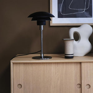 A black table lamp stands on a light oak cabinet; next to it, a ceramic vase and a white abstract sculpture stand in front of a framed black-and-white graphic poster on a brown wall.