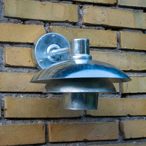 Metal outdoor lamp mounted on a yellow brick wall; round, glossy shade reflects light, short arm and round wall bracket secures the lamp, surrounded by horizontally laid yellow bricks.