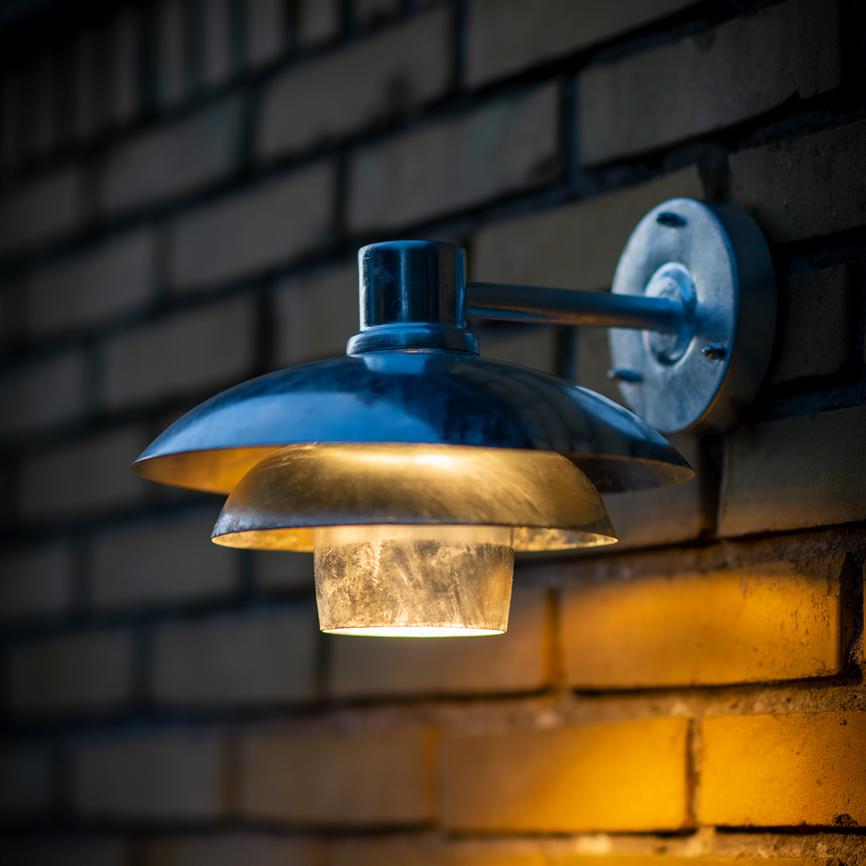 Metal wall lamp with curved lampshade, warm downwards; mounted on a dark brick wall, casts golden light and creates an evening atmosphere.