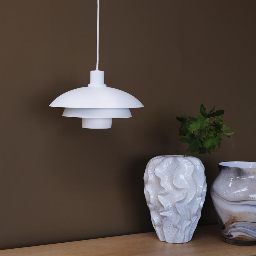 White pendant lamp hangs above a light wooden table; next to it is a rippled white vase with green leaves and a marbled brown-white vase in front of a brown wall.
