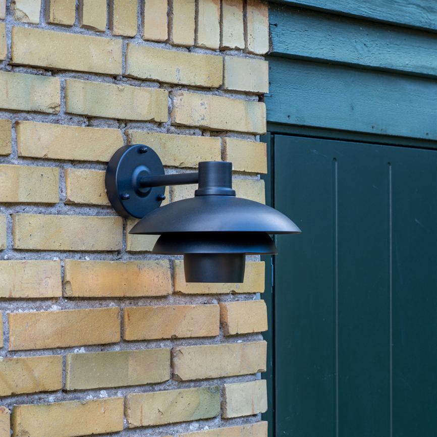 Black metal wall lamp is mounted on a yellow brick wall, points downwards and hangs from a round wall bracket; it sits next to a dark green wooden door and wooden panelling.