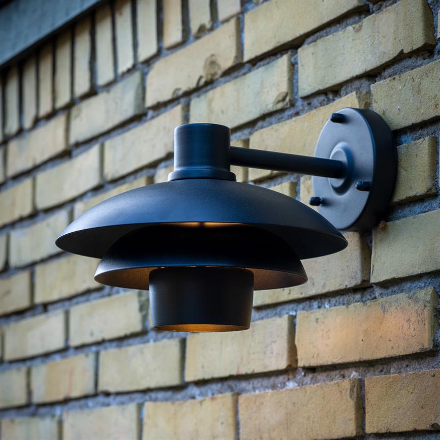 A black, two-piece metal wall lamp emits a warm, downward glow; it is mounted on a round wall bracket and sits on a tan brick facade.
