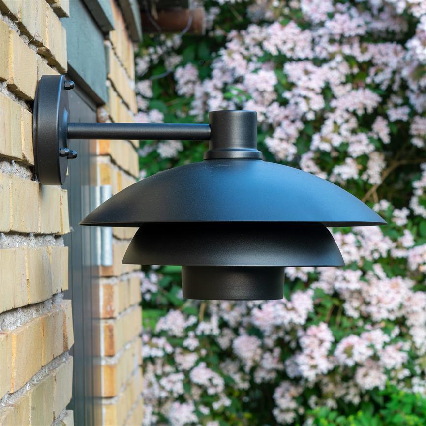 Black, matte, three-layer outdoor wall lamp mounted on a yellow brick wall; a round arm holds the lamp out from the facade, with green bush covered in pink flowers in the background.