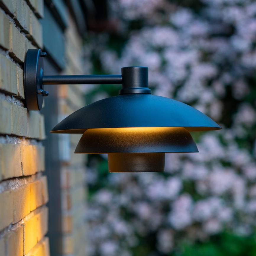 A black metal outdoor wall lamp emits warm golden light, mounted on a pale yellow brick wall with a blurred bush with pink flowers in the background.
