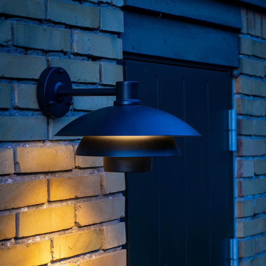 Black, round wall lamp casts warm light downwards and illuminates yellow bricks; mounted on a short arm next to a dark door in dim, cool evening lighting.