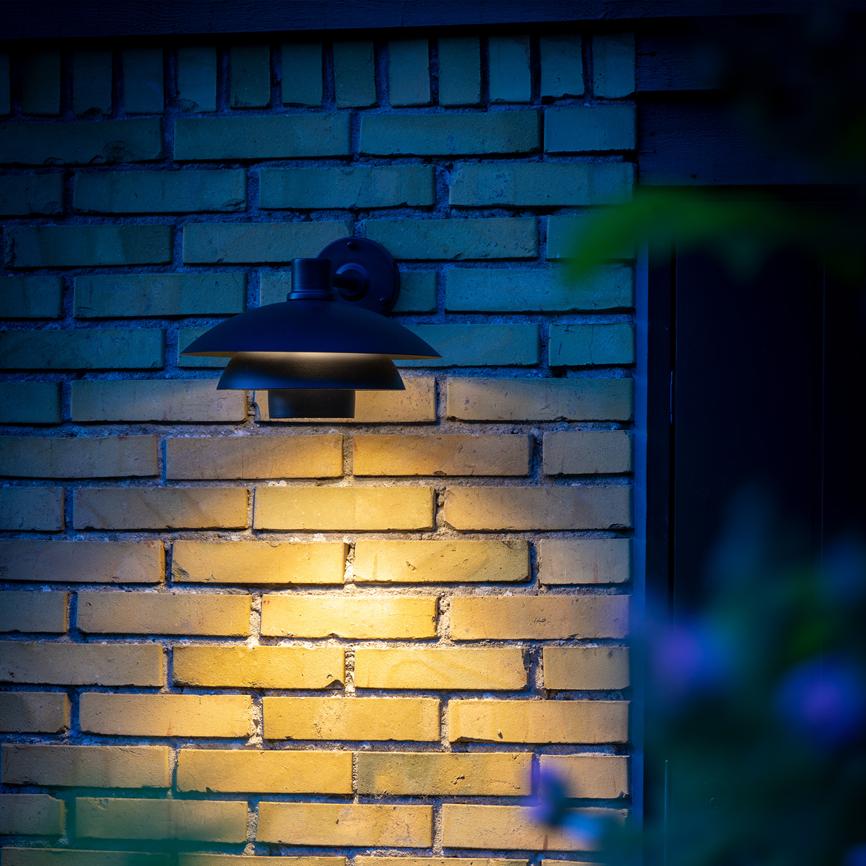 Outdoor wall lamp shines warm downwards against tawny bricks, creating a soft yellow glow; the surroundings are blue-toned in the evening light with blurred leaves in the foreground.