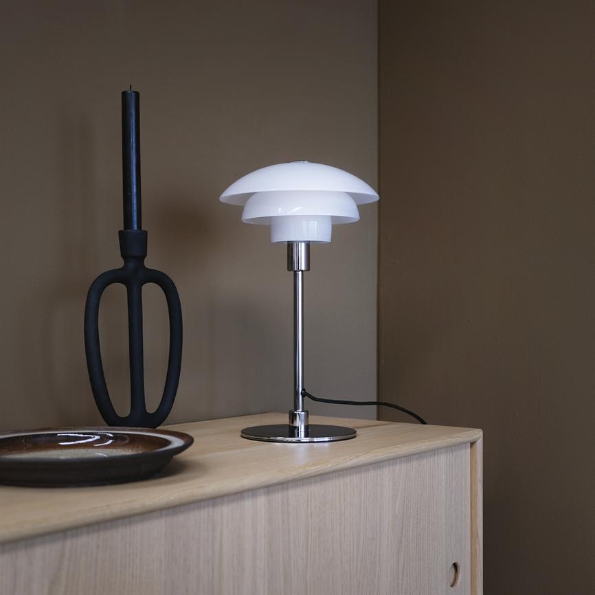 A white shade-shaped table lamp stands on a light wooden cabinet; next to a black candlestick and a brown ceramic bowl, in front of a brown wall.