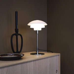 A table lamp shines softly on a light wooden chest of drawers in front of a dark wall; next to it is a black candlestick and a dark, round ceramic bowl.