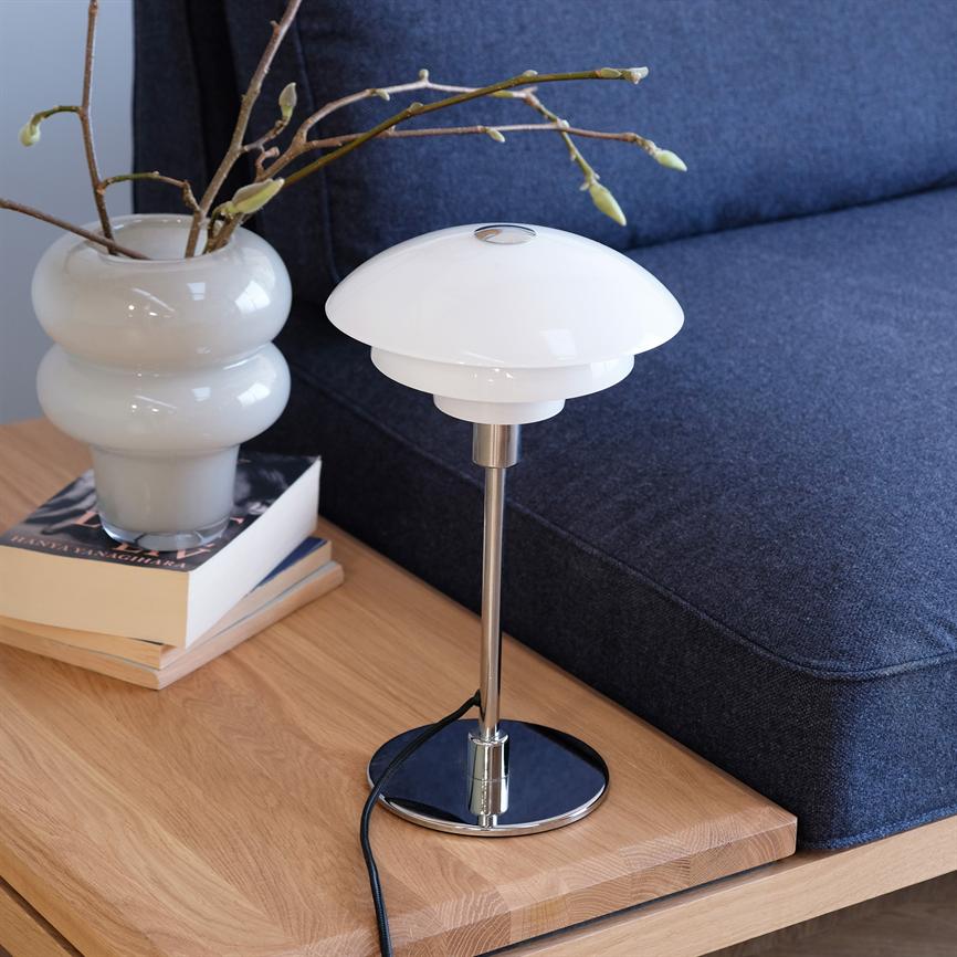 Table lamp is switched off on a light wooden table; white glass shade and chrome foot, next to a white vase with branches and a blue sofa in the background.