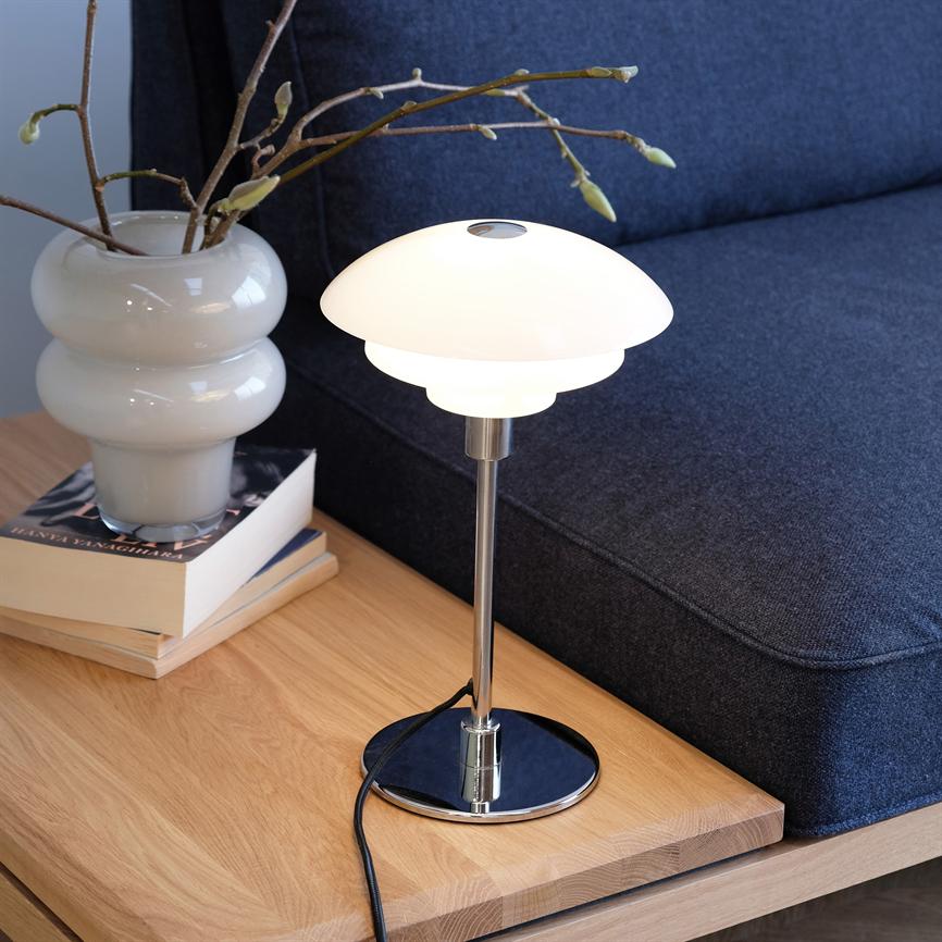A modern white table lamp shines softly on a wooden table; it stands next to a white vase with buds and a stack of books in front of a dark gray sofa in the living room.