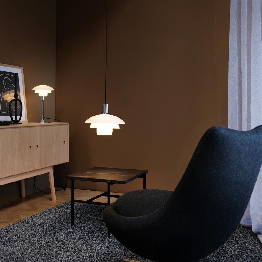 Hanging lamp shines softly above a small coffee table; surrounded by a black, curved armchair, gray carpet, light wooden cabinet and dark wall by a light curtain.