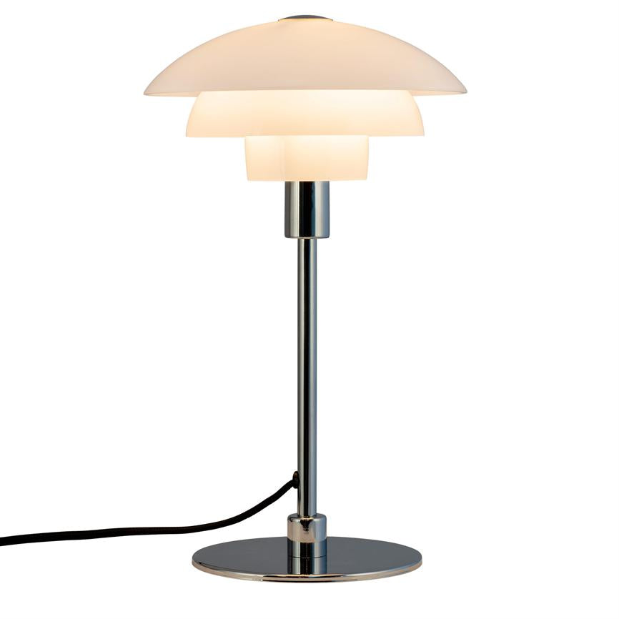 Table lamp with white, three-part shade glows softly; mounted on slim chrome rod and round chrome base, black cord extending to left on neutral white background.