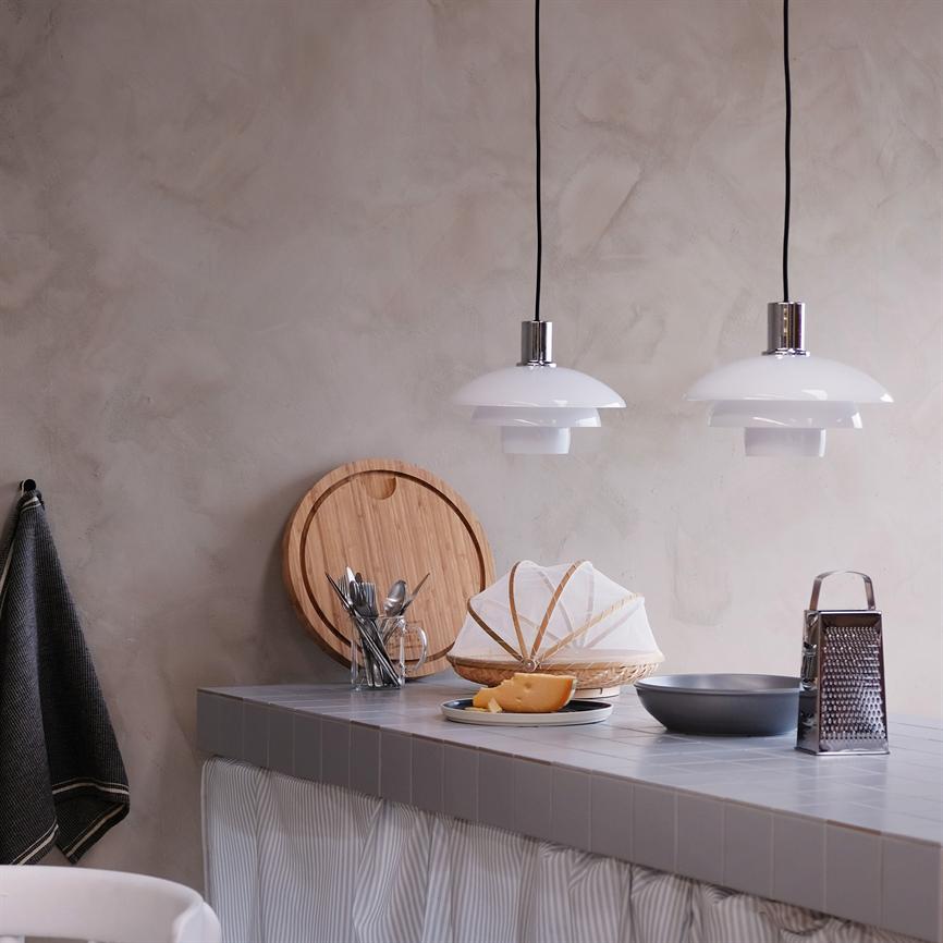 Two white hanging lamps hang above a tiled kitchen table, where a bread basket with fabric netting stands next to pieces of cheese, grater, cutting board and a glass with cutlery in front of a plastered wall.