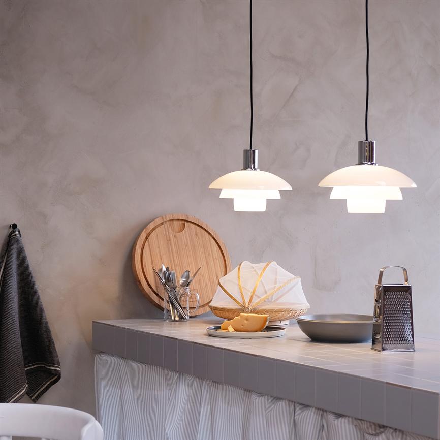 Two white pendant lamps shine above a kitchen table with a tiled table top, cutting board, glass with cutlery, cheese on a plate covered with netting, grater and gray plastered wall.