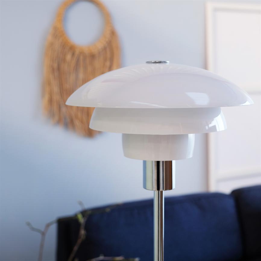 A white glossy table lamp sits unlit on a chrome rod in front of a dark blue sofa in a bright room with a wicker rattan wall decoration and a slim picture frame on the wall.