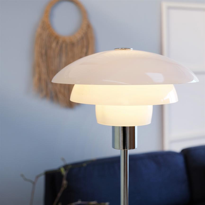 En moderne hvid gulvlampe lyser blødt; den står på kromstativ foran en mørkeblå sofa, med en lyseblå væg bagved dekoreret med flettet vægdekoration og billedramme.