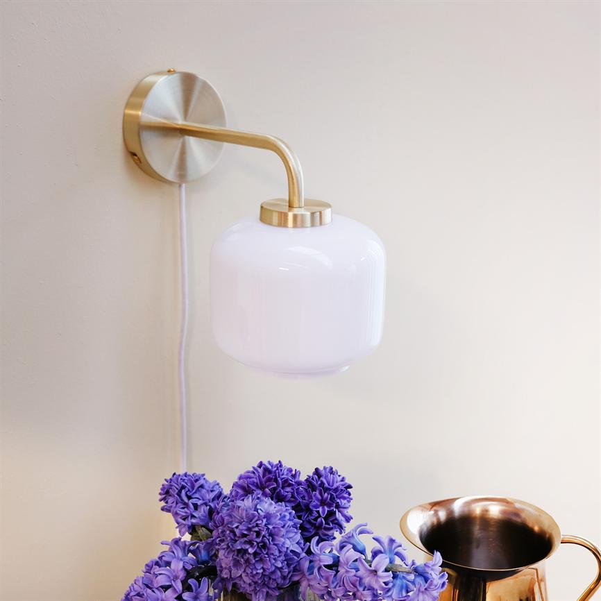Wall lamp in brass with round, white glass shade mounted on a light wall; below it a bouquet of purple hyacinths and a copper jug ​​on a flat.