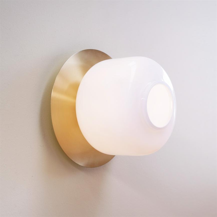 Wall lamp with round, opal glass dome glows softly; mounted on a round, matt brass plate against a light gray wall.