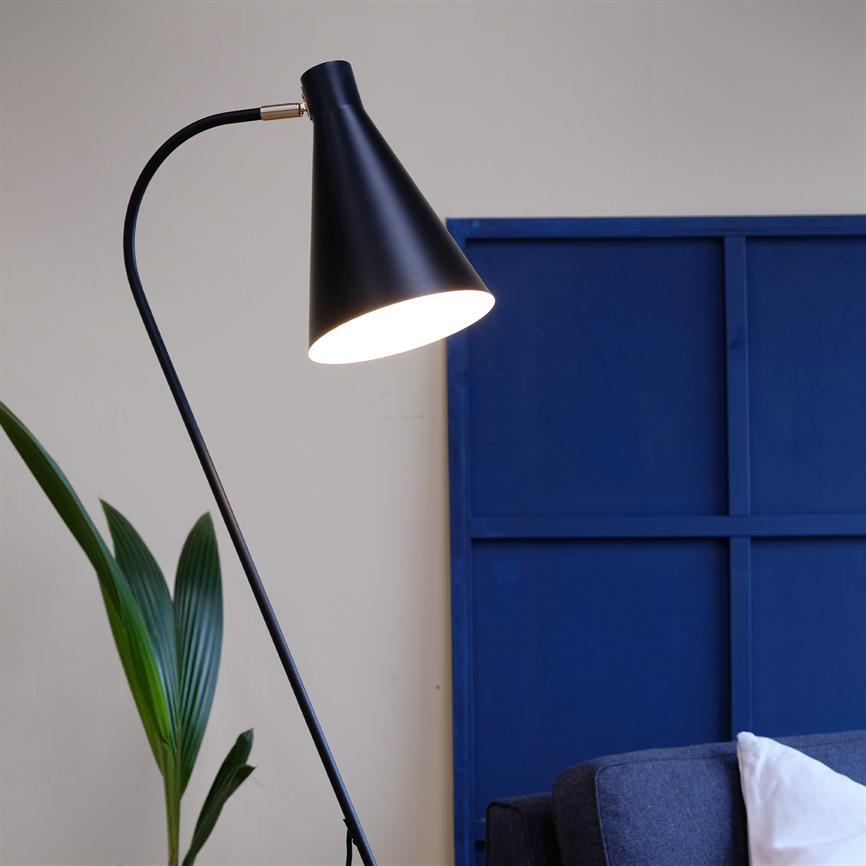 Black, cone-shaped floor lamp shines downwards next to a dark gray sofa; standing in front of a large blue panel and by a green houseplant in a bright, neutral room.