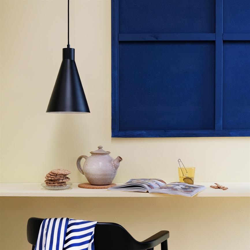Teapot stands on wall-hung shelf, surrounded by stack of biscuits, open magazine and glass of tea; black pendant lamp hangs above, blue frame on yellow wall in kitchen corner.
